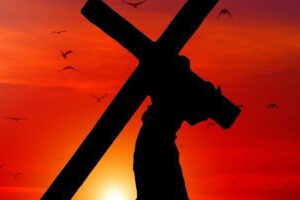 Man going up a hill carrying a cross, silhouetted against a sunset