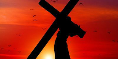 Man going up a hill carrying a cross, silhouetted against a sunset