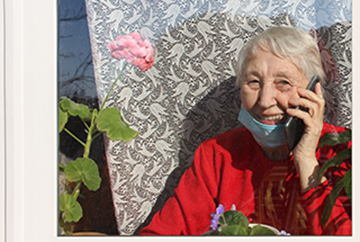 Old woman wearing protective medical mask sitting near the window at home with mobile phone