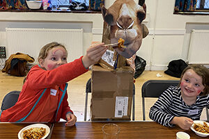 Two girls sit either side of a cut-out picture of a camel at Messy Church, St Barnabas, Dulwich