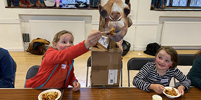 Two girls sit either side of a cut-out picture of a camel at Messy Church, St Barnabas, Dulwich