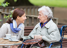 Over 90-year-old woman in a wheelchair and a home caregiver drinking cup of coffee outdoors in a park